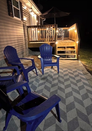An outdoor patio with two blue Adirondack chairs, a wooden deck, and a patterned rug.