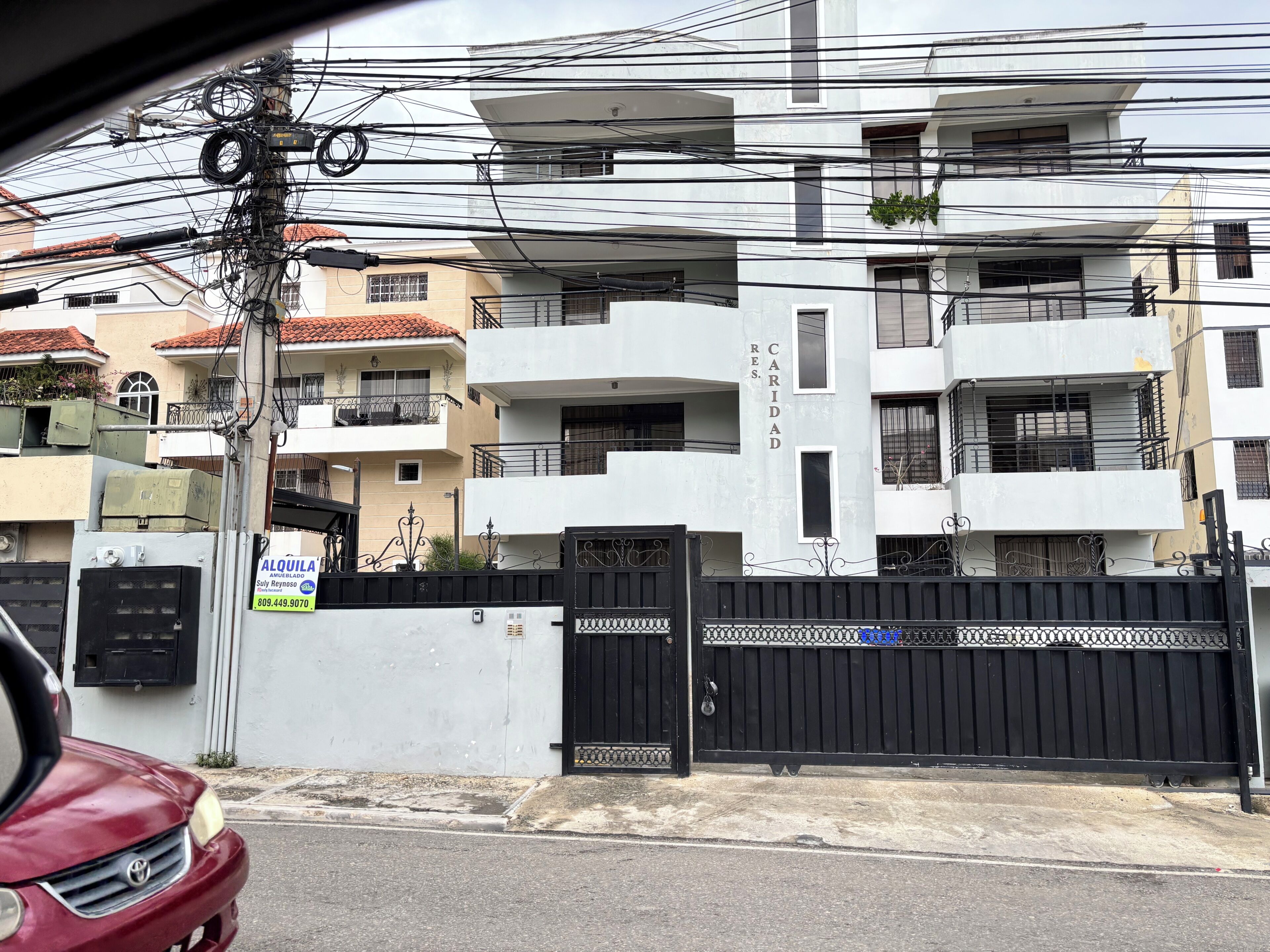 Um edifício residencial de vários andares com varandas, entrada com portão e infraestrutura elétrica visível.