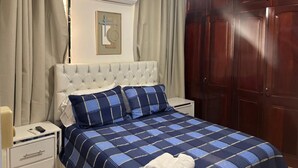 Una camera da letto con testiera trapuntata, un letto con copripiumino blu e bianco a quadri, un comodino bianco con un telecomando e una ventola a soffitto.