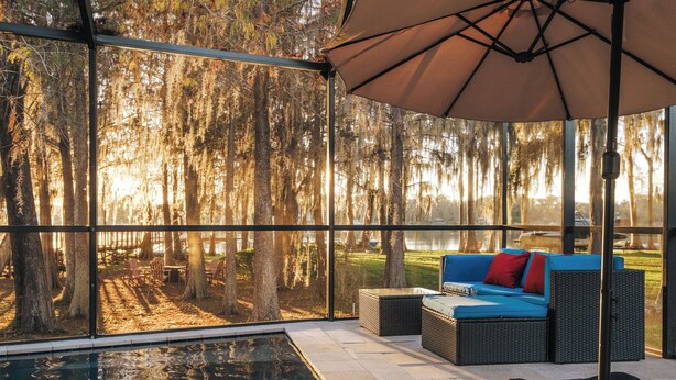 An indoor pool area with a view of a forest, a blue sofa with red cushions, and a large umbrella. - Luxury Lakefront Retreat w Pool, Balcony, Firepit, Dock, Orlando, FL Sleeps 12 (Orlando)