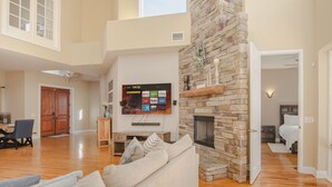 A spacious living room with a stone fireplace, a flat-screen TV mounted on the wall, and a dining area with wooden flooring. - Luxury Lakefront Retreat w Pool, Balcony, Firepit, Dock, Orlando, FL Sleeps 12 (Orlando)