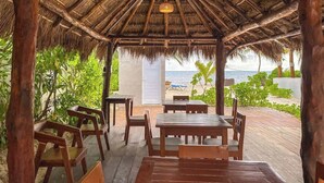 Een houten terras met tafels en stoelen, een rieten dak en uitzicht op de oceaan.