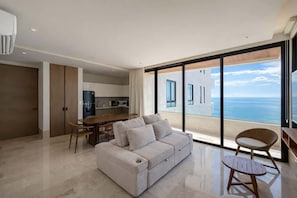 A modern living room with a sofa, a dining table, and chairs. There is a large window offering a view of the sea.