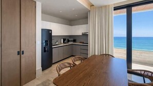 Una cocina moderna con refrigerador, microondas y cafetera, una mesa de comedor de madera con sillas y un amplio ventanal con vista al océano.