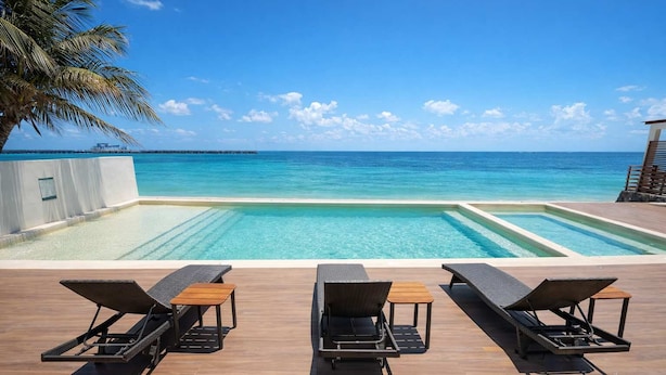 A pool with clear blue water, surrounded by lounge chairs and a wooden deck, overlooking a calm sea. - Beachfront 3BR Puerto Morelos - Rooftop Pool (Puerto Morelos)