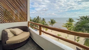 En balkon med en fletsofa, gelænder i træ og udsigt til stranden med palmer.