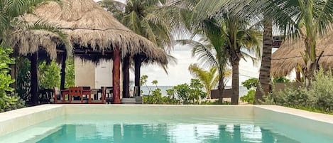 A clear swimming pool surrounded by palm trees and a thatched-roof structure.