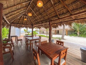 Een overdekt terras buiten met houten tafels en stoelen, een rieten dak en uitzicht op het strand.