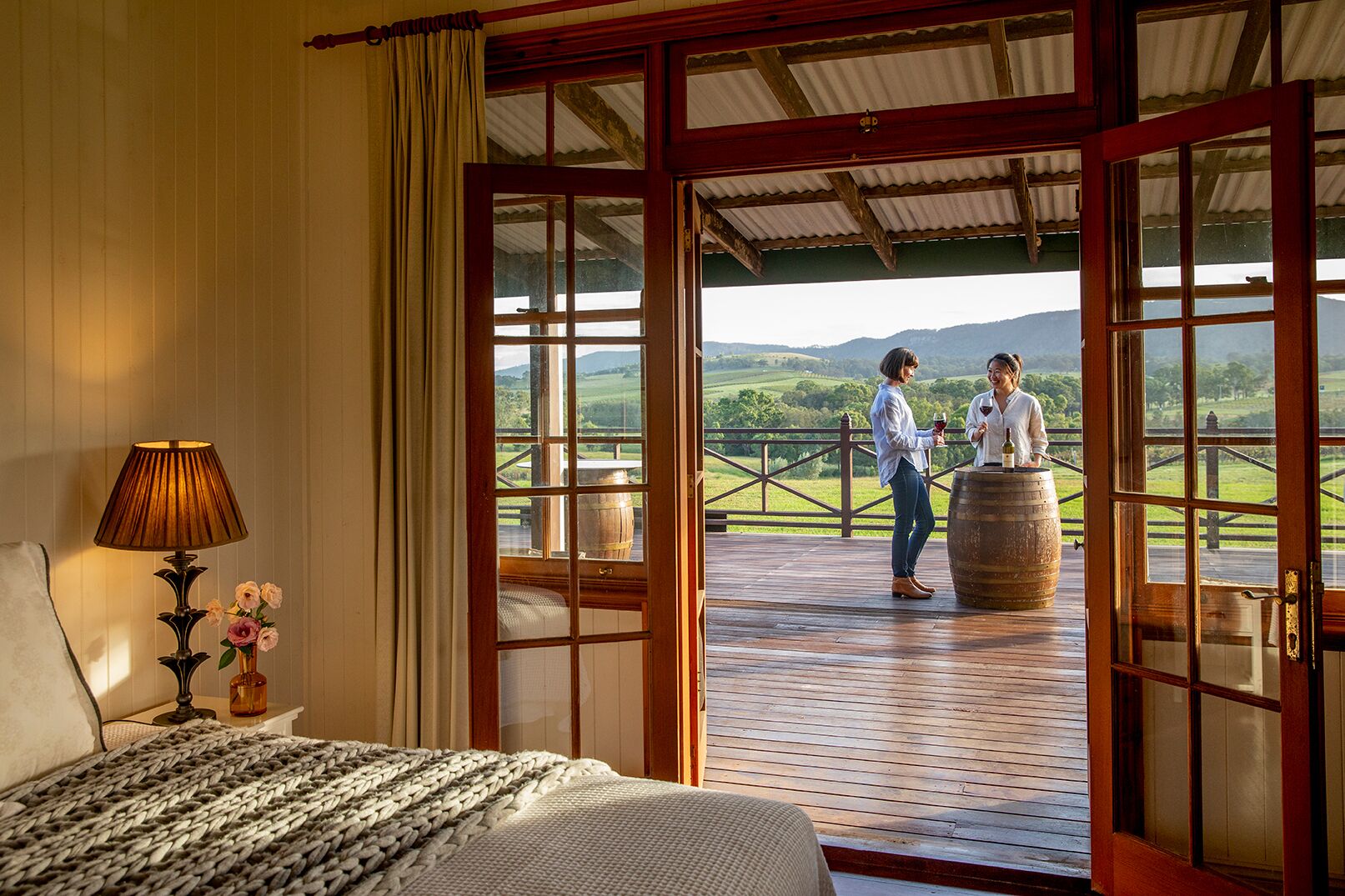 Una habitación con cama, lámpara y vista a un balcón donde dos personas sostienen copas de vino.