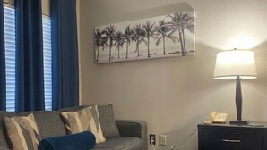 A living room with a grey sofa, blue cushions, a blue ottoman, a painting of palm trees, a lamp, and a telephone on a black side table. - Cozy Studio near Universal (Orlando)