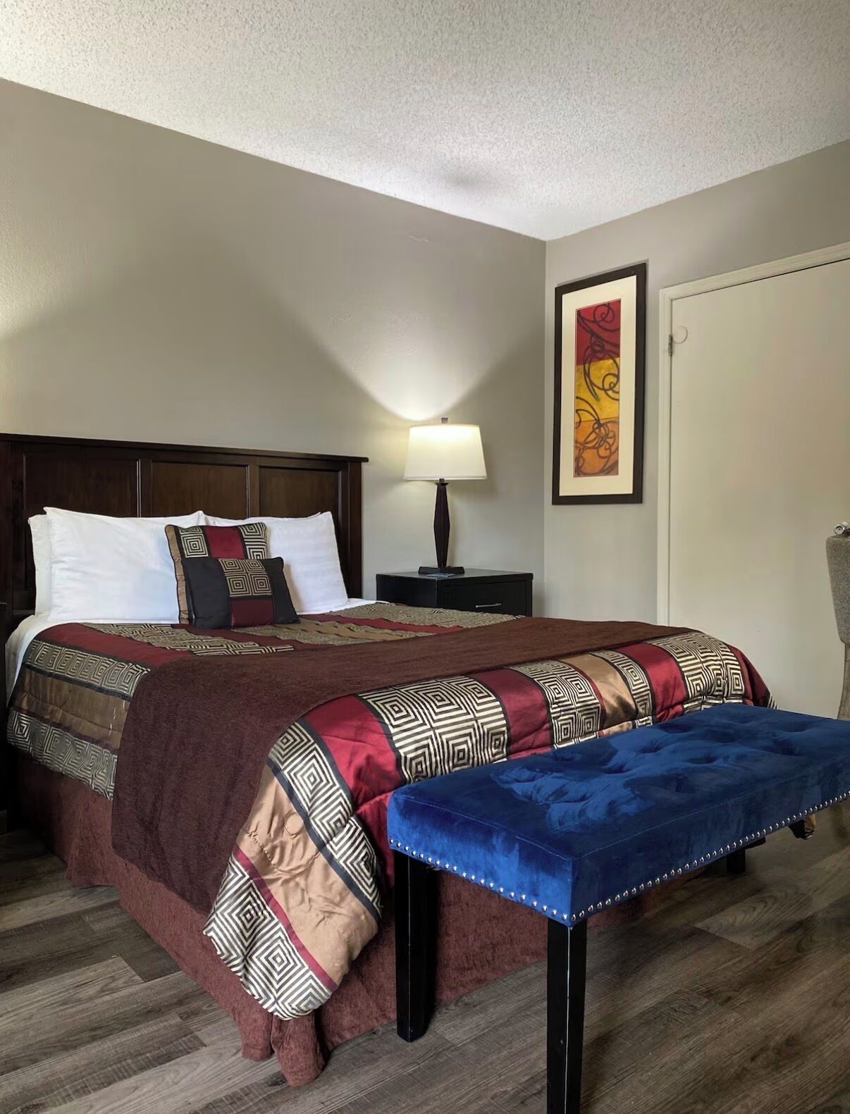 A hotel room with a large bed, a bedside table with a lamp, a framed artwork on the wall, and a blue bench.