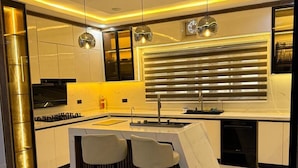 Een moderne keuken met een centraal eiland, twee barkrukken, hanglampen en een raam met jaloezieën.