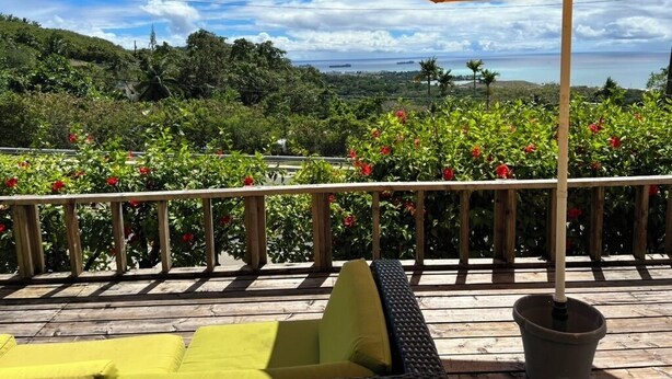 Una terraza de madera con cojines amarillos y un paraguas naranja, con vistas a un jardín exuberante y una lejana vista del mar.