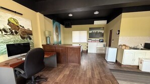 Una habitación amplia con piso de madera, un escritorio con computadora, una silla, un área de cocina con refrigerador y una ventana con persianas.