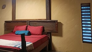 Een slaapkamer met een houten bed, rode en blauwe kussens, een houten hoofdbord, een plafondventilator en een raam met jaloezieën.