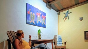 Iemand zit op een houten stoel in een kamer met een schilderij aan de muur en een klein tafeltje met een fles.