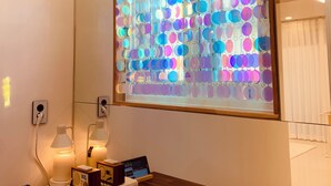 Una camera d'albergo con un'ampia finestra che mostra un motivo circolare dai colori vivaci, un tavolo di legno con diversi dispositivi elettronici e un cartello a parete con la scritta "WINSOME".