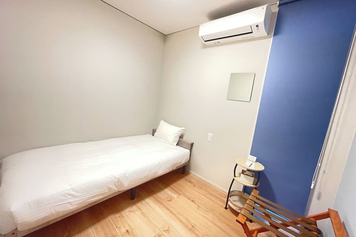 Ein kleines Zimmer mit einem Einzelbett, Holzboden, einem weißen Bett, einem kleinen Tisch und einer an der Wand befestigten Klimaanlage.