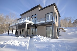 A modern two-story house with a balcony, large windows, and a snowy landscape. - Modern Chalet in Bel Air Resort Tremblant (La Conception)