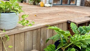 Um deck de madeira com plantas em vasos e uma casa com janelas amplas e telhado inclinado.