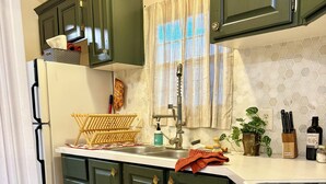 Uma cozinha com armários verde escuro, bancada branca, pia de aço inoxidável e micro-ondas. Há um tapete estampado no chão.