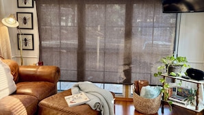 Uma sala de estar aconchegante com um sofá de couro marrom, uma janela grande com vista para árvores, uma TV de tela plana e uma estante com livros e uma planta.
