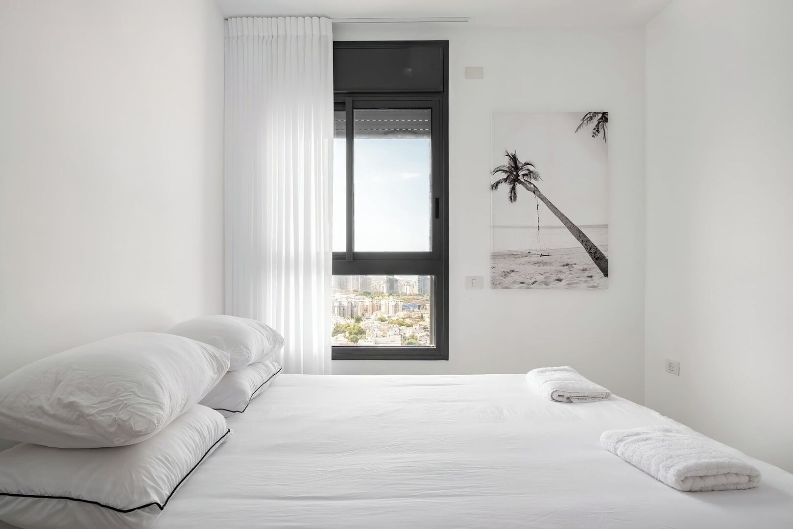 A minimalist bedroom with a large bed, white bedding, a view of a cityscape through a window, and a beach-themed wall art.