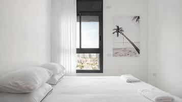 A minimalist bedroom with a large bed, white bedding, a view of a cityscape through a window, and a beach-themed wall art.