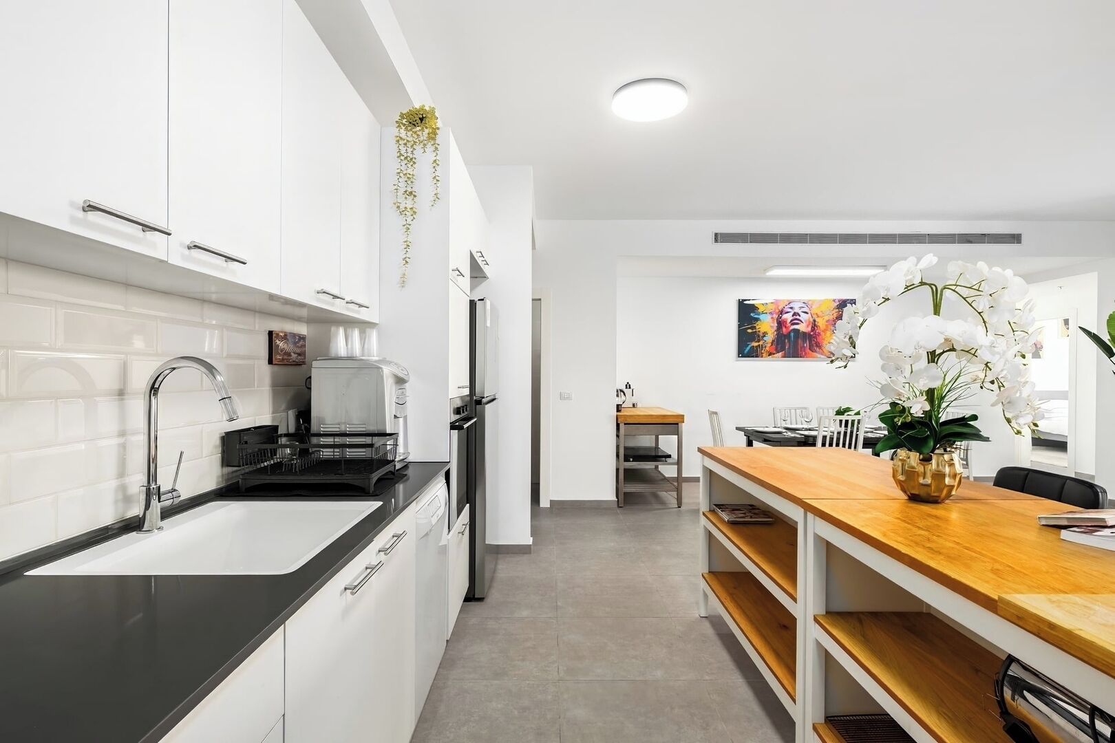 A modern kitchen with white cabinets, a black countertop, and stainless steel appliances. There is a dining area with a wooden table and chairs, and a painting on the wall.