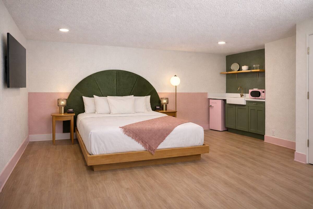 A bedroom with a bed, bedside tables, a lamp, a small kitchenette, and a TV mounted on the wall.