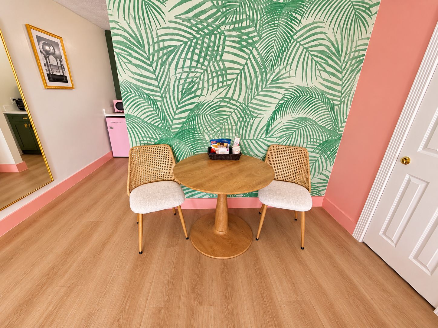 A room with a round wooden table, two wicker chairs, a wall with a tropical leaf pattern, a door, and a mirror reflecting part of the room.