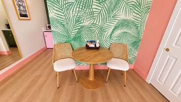 A room with a round wooden table, two wicker chairs, a wall with a tropical leaf pattern, a door, and a mirror reflecting part of the room.