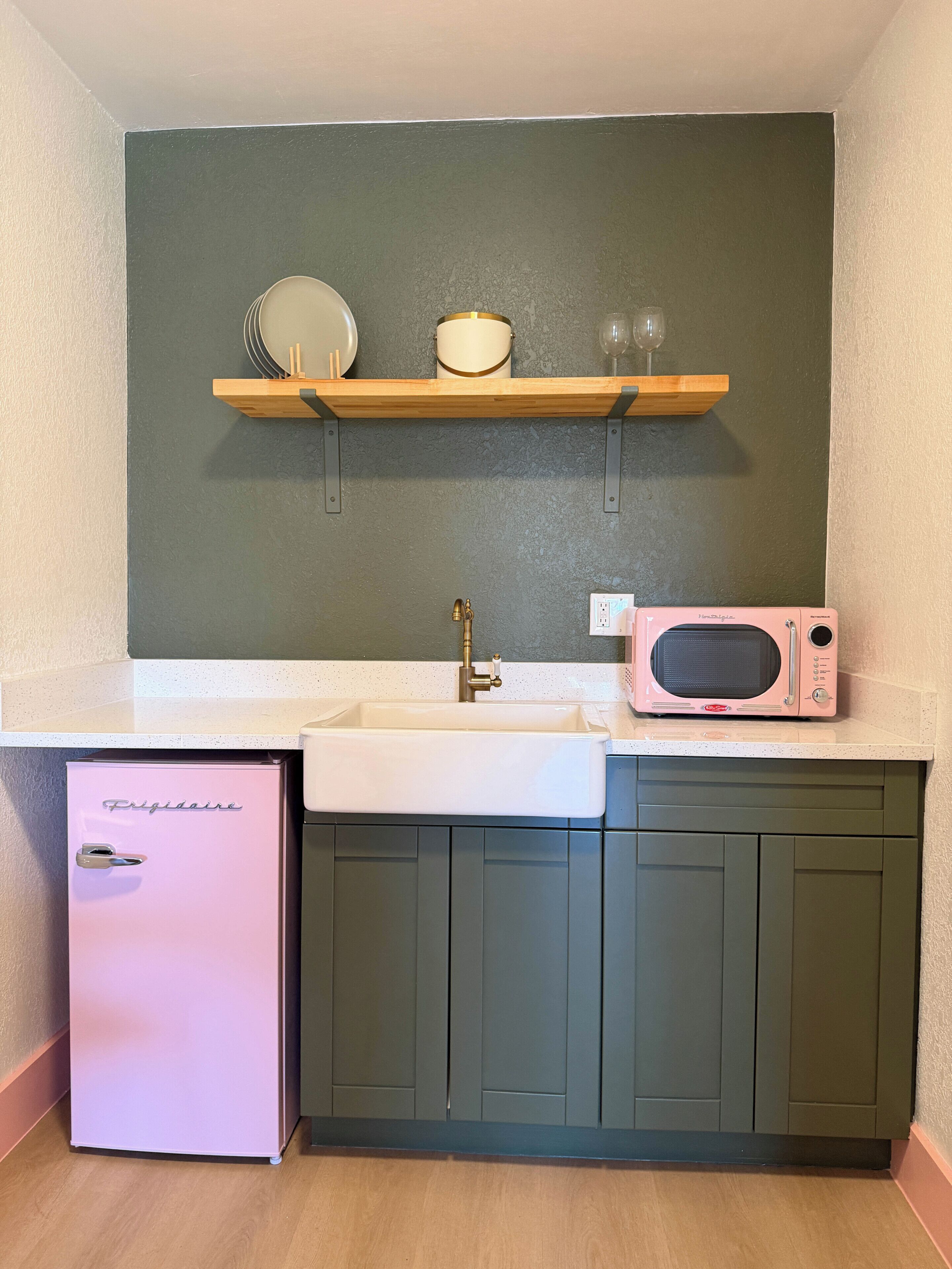 Eine kompakte Küche mit einem weißen Spülbecken, einem pinkfarbenen Frigidaire-Kühlschrank, einer Mikrowelle und einem Holzregal über dem Spülbecken.