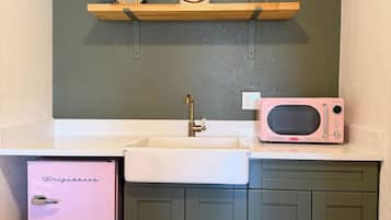 Una cocina compacta con un lavabo blanco, un refrigerador Frigidaire rosa, un microondas y un estante de madera sobre el lavabo.