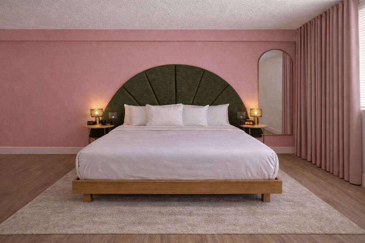 A bedroom with a large bed, two bedside tables with lamps, a large mirror, and a pink wall.