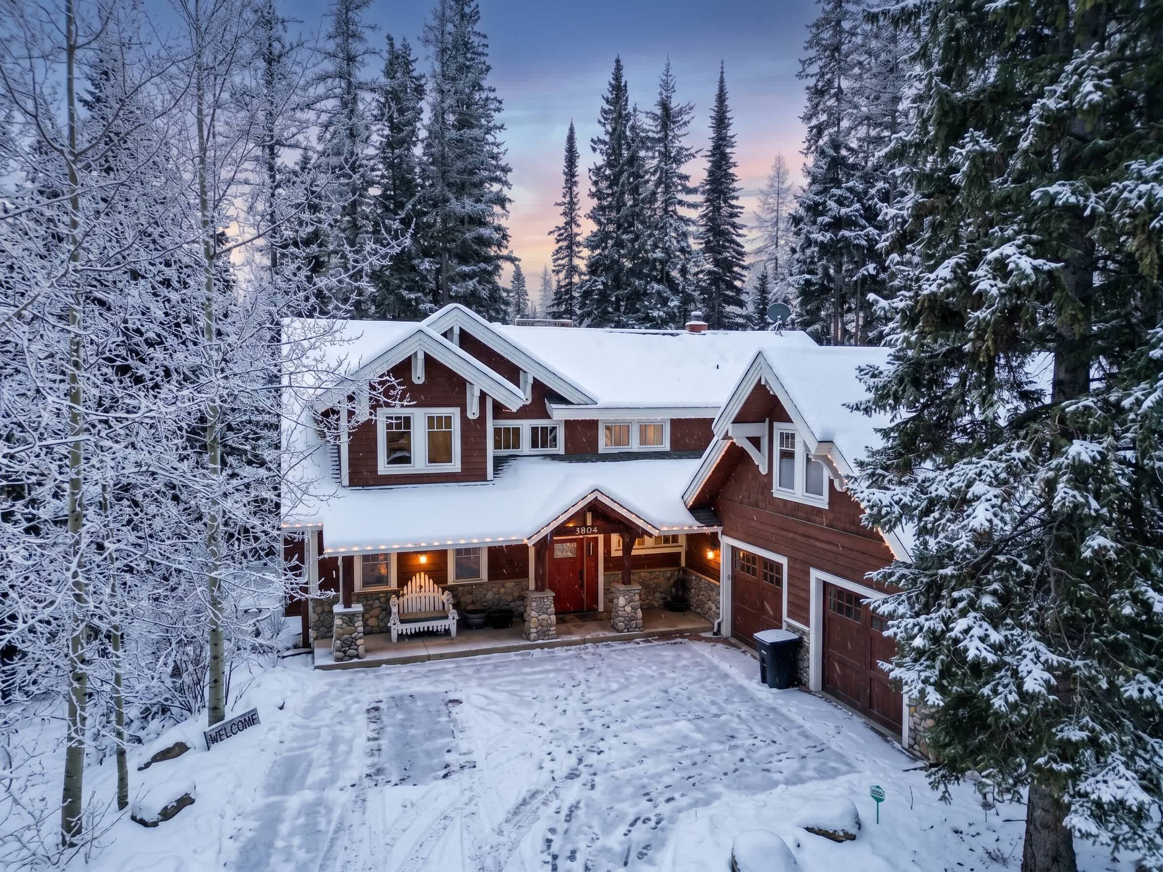 A two-story house with a snow-covered roof and a stone foundation, surrounded by tall evergreen trees.