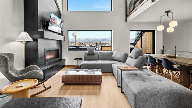 A modern living room with a fireplace, a large window with a city view, a dining area, and a loft space. - Downtown Whitefish Retreat – Hot Tub • Mountain Views • 3 Bedrooms + sleeping Loft (Whitefish)