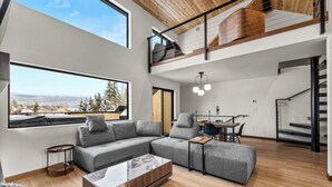 A spacious living area with a large sectional sofa, a glass coffee table, and a dining table with chairs. There is a staircase leading to an upper level, and large windows offering a view of the outdoors. - Downtown Whitefish Retreat – Hot Tub • Mountain Views • 3 Bedrooms + sleeping Loft (Whitefish)