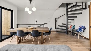 A modern dining area with a wooden table and black chairs, a staircase with a black railing, and a view of the outside through a large window. - Downtown Whitefish Retreat – Hot Tub • Mountain Views • 3 Bedrooms + sleeping Loft (Whitefish)