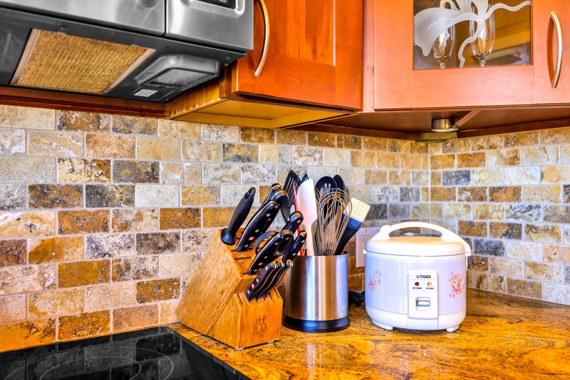 A kitchen with a stone backsplash, a wooden cutting board, a knife block, a stainless steel pot, and a white rice cooker.