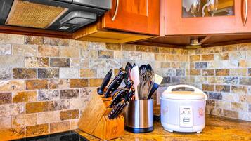A kitchen with a stone backsplash, a wooden cutting board, a knife block, a stainless steel pot, and a white rice cooker.