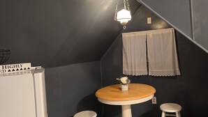 Una habitación pequeña con decoración oscura, una mesa redonda de madera, dos taburetes blancos, un colgante de luz y un pequeño estante en la pared con un jarrón de flores.
