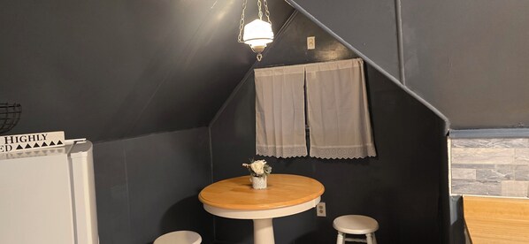A small, dark-themed room with a round wooden table, two white stools, a hanging light fixture, and a small wall-mounted shelf with a vase of flowers.