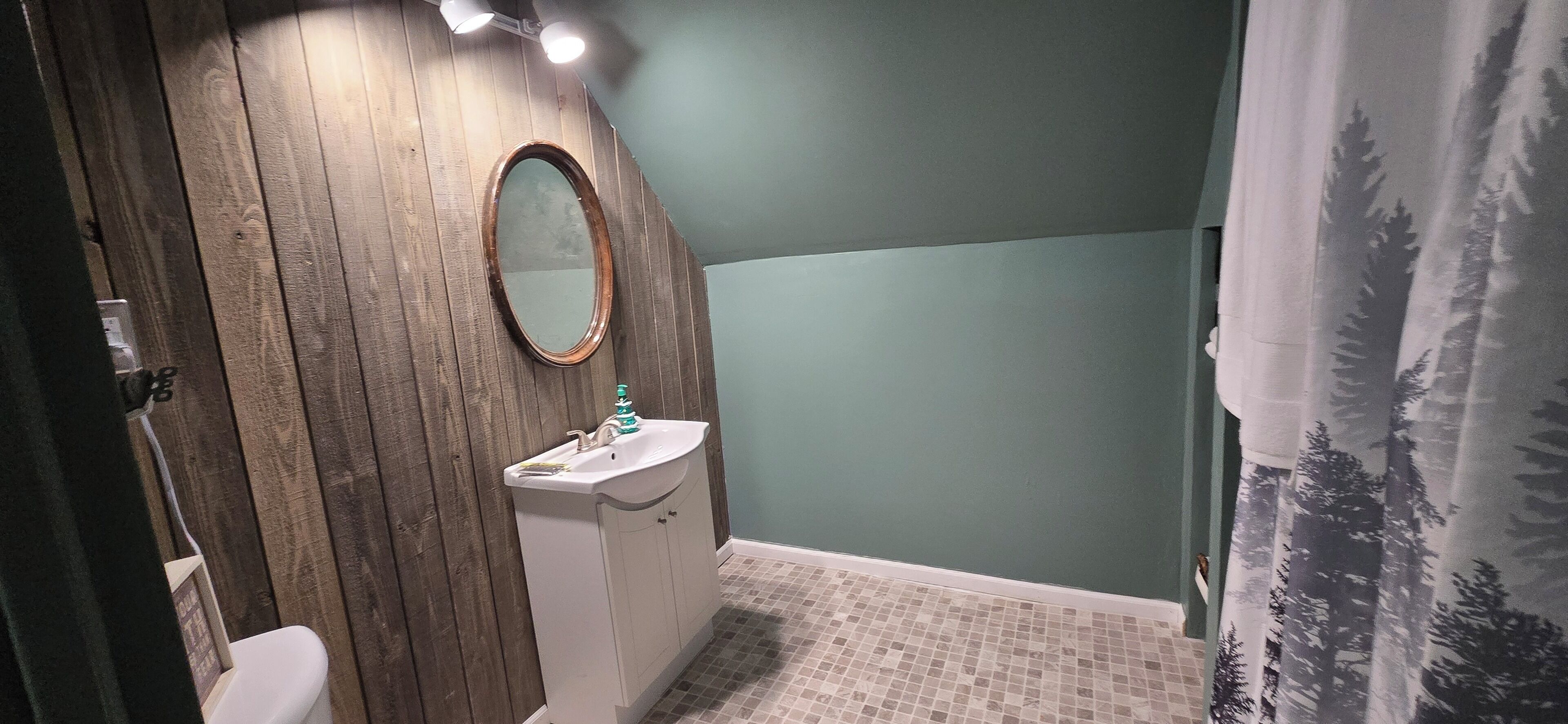 Un baño con tocador de madera, lavamanos blanco, espejo y cortina de ducha con estampado de bosque.