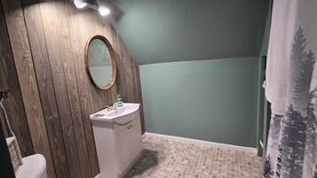 Un baño con tocador de madera, lavamanos blanco, espejo y cortina de ducha con estampado de bosque.