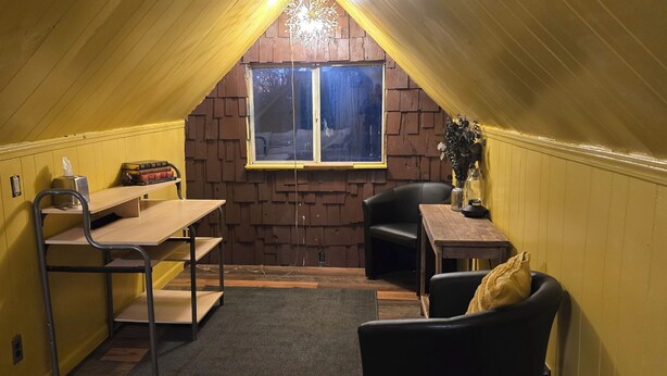 Una habitación acogedora en el ático con techo inclinado, una pequeña mesa con una silla, una ventana con vistas y una estantería con libros y dispensador de pañuelos.