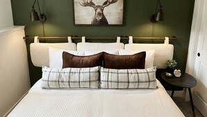 Un dormitorio con cama, almohadas, cabecera, un cuadro colgado en la pared, lámparas de noche y una mesita de noche con un reloj.
