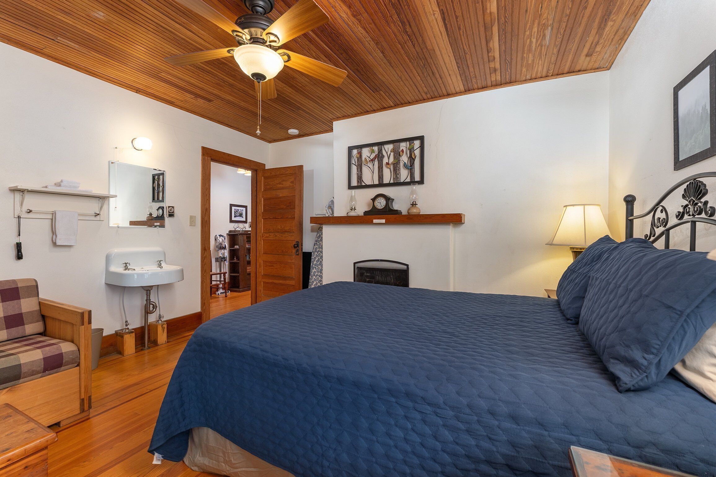 Un dormitorio con techo de madera, una cama con colcha azul, una mesita de noche con lámpara, un estante con adornos y una puerta que da a otra habitación.