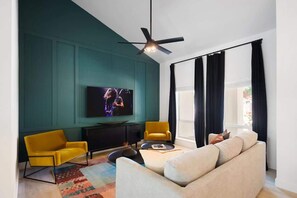 Ein modernes Wohnzimmer mit Türkiswänden, einem Flachbildfernseher, gelben Sesseln, einem beigen Sofa und einem Deckenventilator.