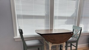 A dining table with two chairs, a window with blinds, and wooden flooring.